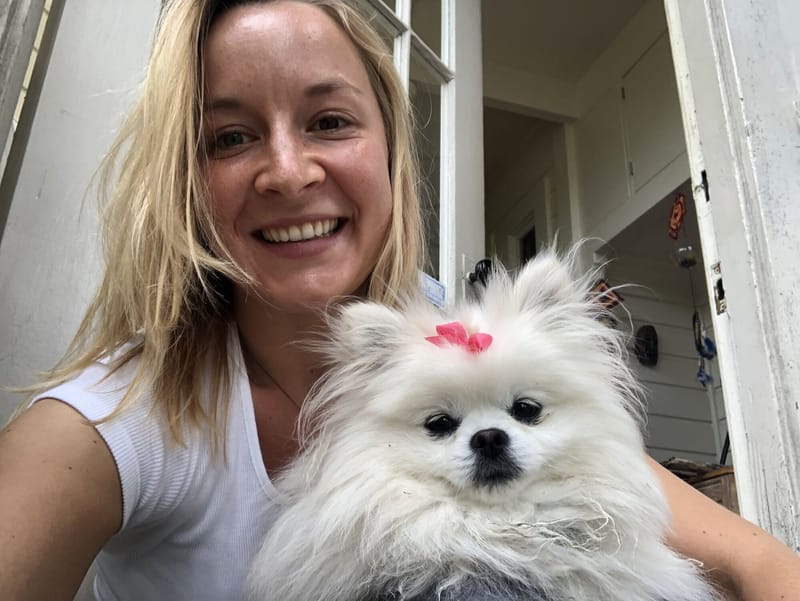 JJ smiling in a doorway, holding a fluffy white Pomeranian with a pink bow