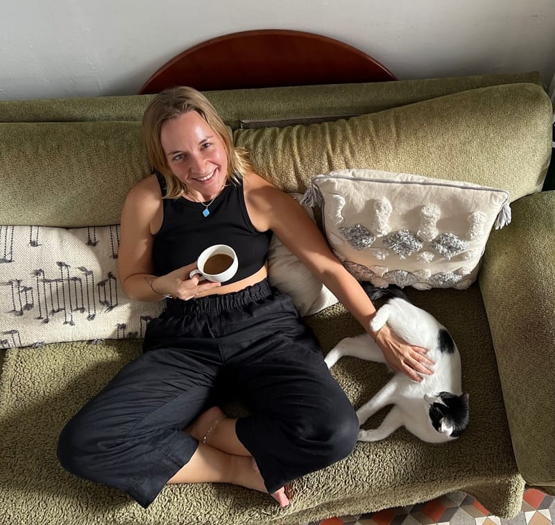 JJ sitting cross-legged on a green sofa with a coffee, stroking a black-and-white cat curled up beside her