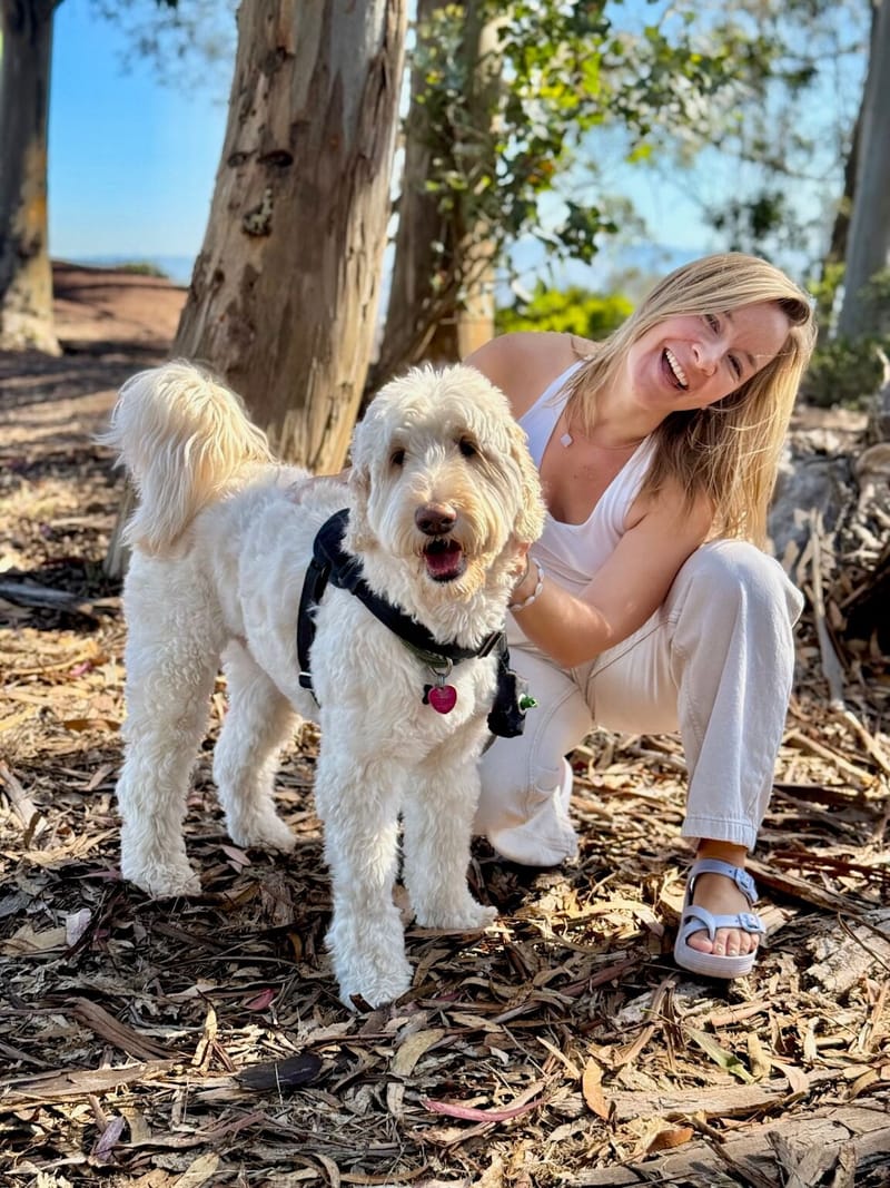 JJ crouching beside a fluffy white doodle in a harness, both smiling, under eucalyptus trees